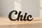 Black wooden decor word 'Chic' on wooden surface with soft shadows in natural light