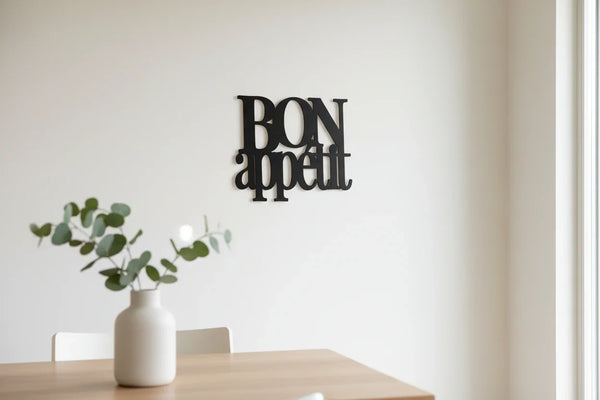 Minimalist kitchen decor with black wooden 'Bon Appetit' wall sign and white vase with green leaves on wooden table