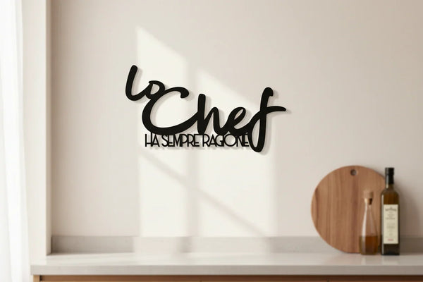 Black wooden wall sign reading "Lo Chef ha sempre ragione" in a minimalist kitchen with cutting board and bottles