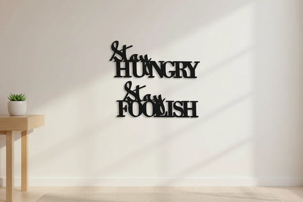 Black wooden wall art saying 'Stay Hungry Stay Foolish' in modern minimalist room with small plant on wooden table