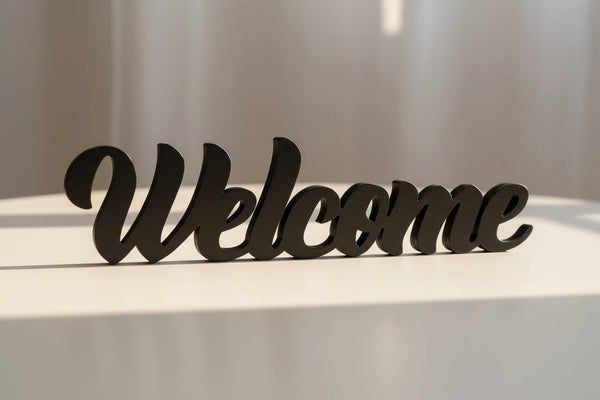 Black wooden decorative sign spelling 'Welcome' on a light surface with soft sunlight, home decor