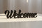 Black wooden decorative sign spelling 'Welcome' on a light surface with soft sunlight, home decor
