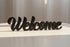 Black wooden decorative sign spelling 'Welcome' on a light surface with soft sunlight, home decor